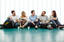 Fünf junge Menschen sitzen nebeneinander an eine Glaswand gelehnt